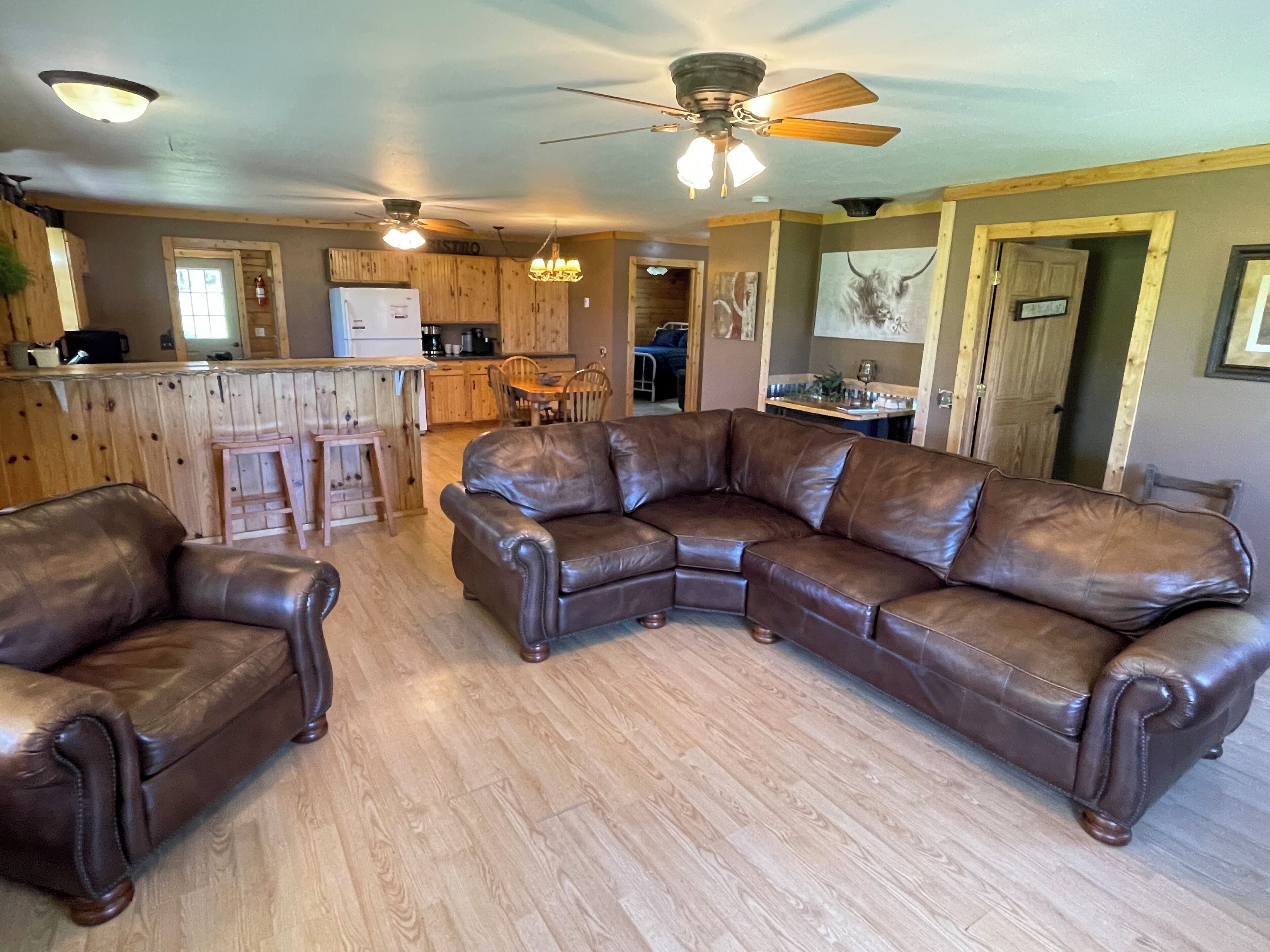 Ein Wohnzimmer mit einem Ecksofa, Holzboden, Deckenventilator und einer Küche im Hintergrund.
