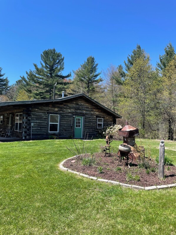 A rustic log cabin with a green door, surrounded by a well-maintained lawn and a garden with a bird bath and decorative rocks.