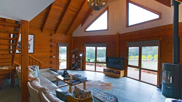 Amplia sala de estar con techo alto, vigas de madera, chimenea, televisor y grandes ventanales con vistas al exterior.