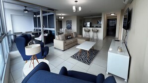 Ein modernes Wohnzimmer mit einer Couch, Sesseln und einem Couchtisch. Es gibt eine Küchenzeile mit Kühlschrank und Mikrowelle. Das Zimmer verfügt über große Fenster und einen Deckenventilator.