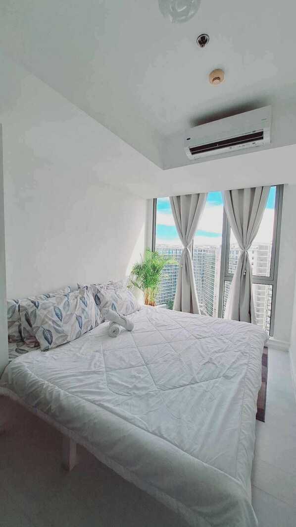 A modern bedroom with a large bed, an air conditioning unit, a potted plant, and a view of the city through large windows.
