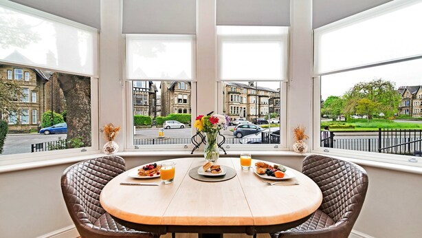 A dining area with a table set for breakfast, large windows with a view of residential buildings, and a vase with flowers. - Apartment in Harrogate Near Turkish Baths (Harrogate)