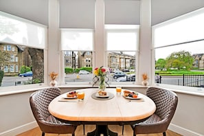 A dining area with a table set for breakfast, large windows with a view of residential buildings, and a vase with flowers. - Apartment in Harrogate Near Turkish Baths (Harrogate)