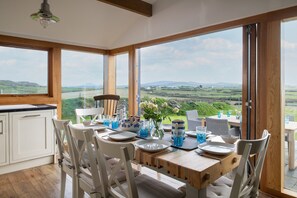 A dining area with a table set for four, large windows offering a view of the countryside, and a wooden ceiling. - Coastal Retreat on Wales Path With Sea Views (Pwllheli)