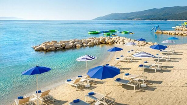 A beach with clear blue water, a rocky breakwater, and several blue umbrellas and sun loungers. - Pleasant Apartment With Pool, Sauna, Hot Tub (Rasa)