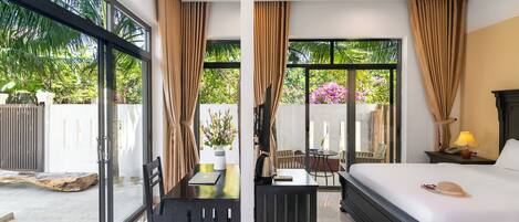 A modern hotel room with a bed, a desk, and a dining table. There are large windows with curtains, a view of the garden outside.