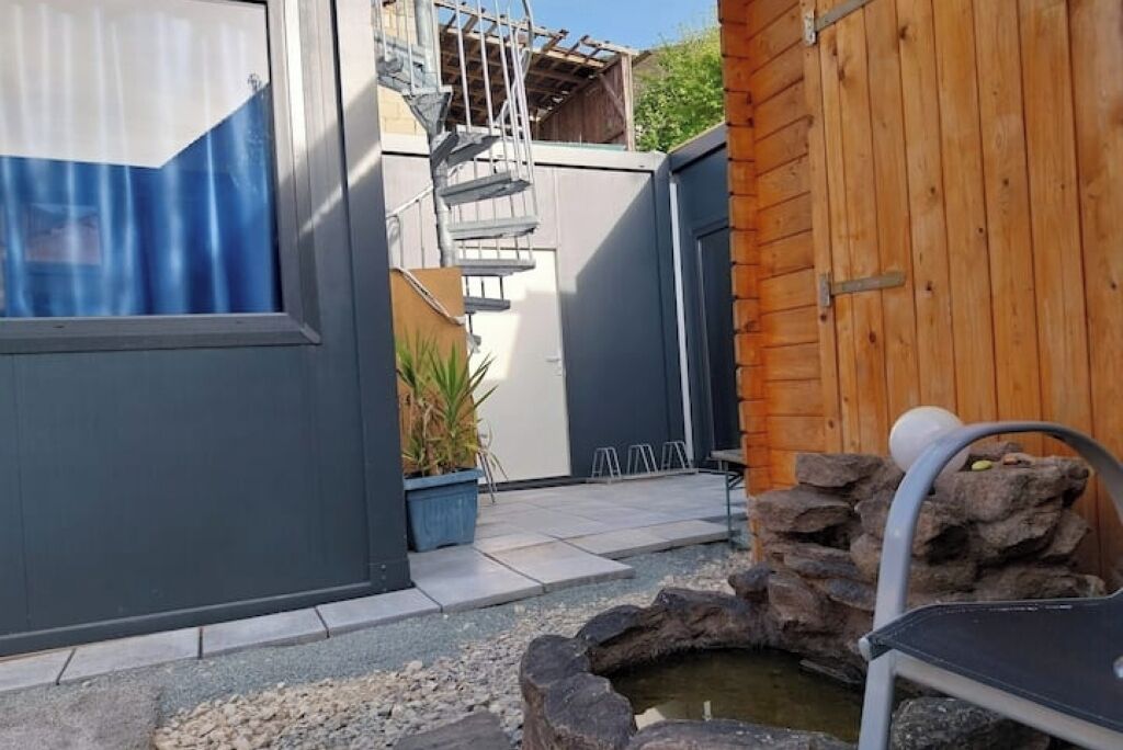 Un edificio moderno con fachada gris, puerta de madera y un pequeño espacio exterior con pared de piedra y fuente de agua.