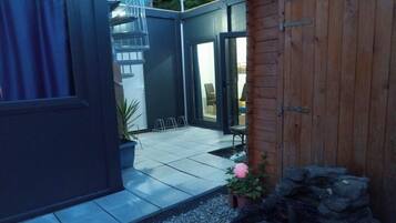 Una zona patio con pavimento piastrellato, recinzione in legno e un piccolo giardino con un fiore rosa.