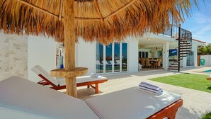 Una villa moderna con piscina, sillas de descanso, zona de comedor y escalera que sube al piso superior.