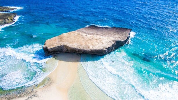Una playa con una gran formación rocosa, agua turquesa cristalina y una orilla arenosa.