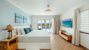 Un dormitorio con una cama grande, un televisor, una mesita de noche de madera y vista al océano a través de la ventana.