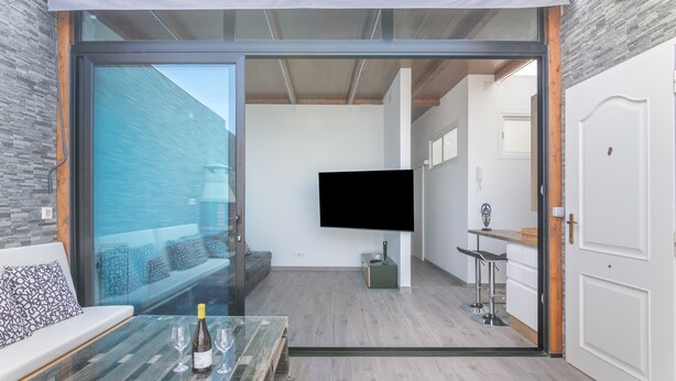 Ein modernes Wohnzimmer mit Glastür zum Balkon, einem Wand-Fernseher, einem Essbereich mit Barhocker und einer Küchenzeile mit Spüle und Arbeitsfläche.