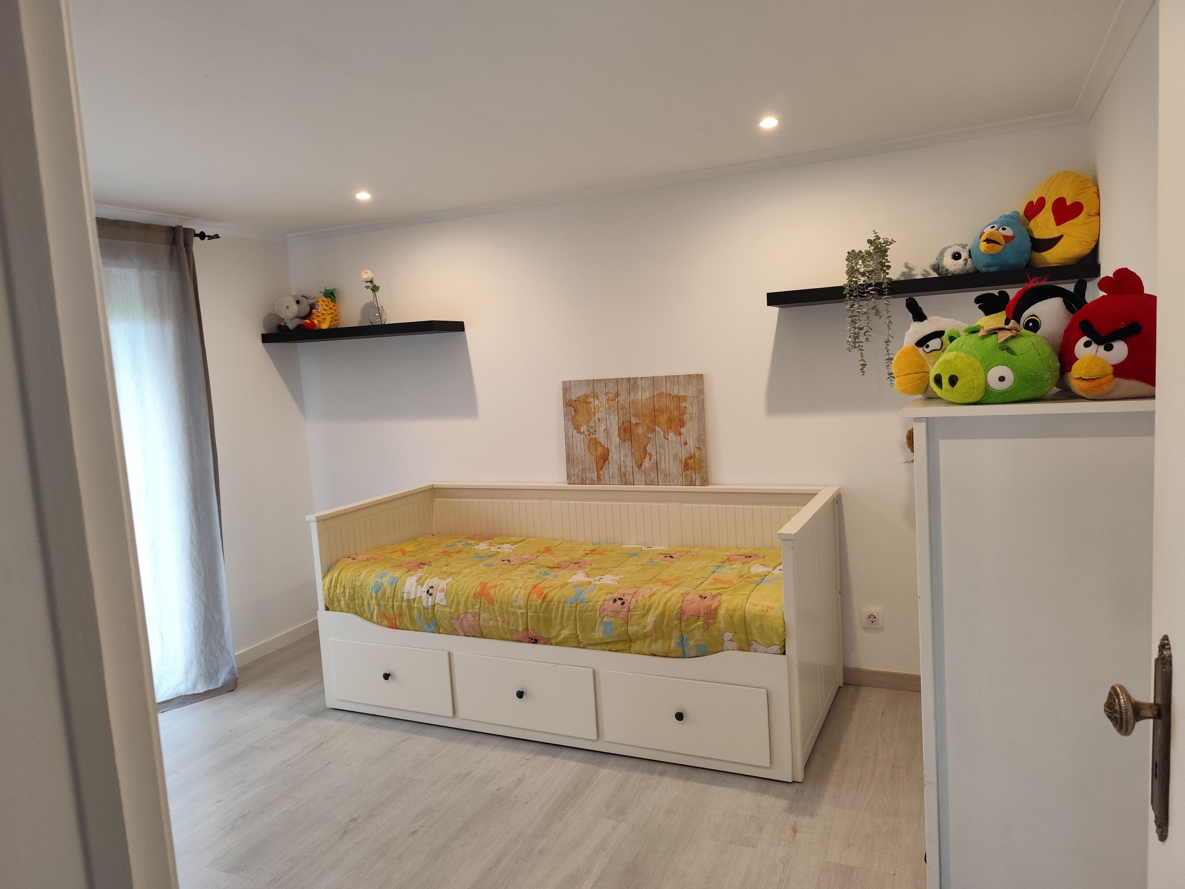Une chambre avec un lit, une commode, une étagère remplie de peluches et une carte du monde accrochée au mur.