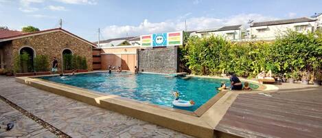 A swimming pool with stone tiles, a wooden deck, and a stone building with arched doorways.