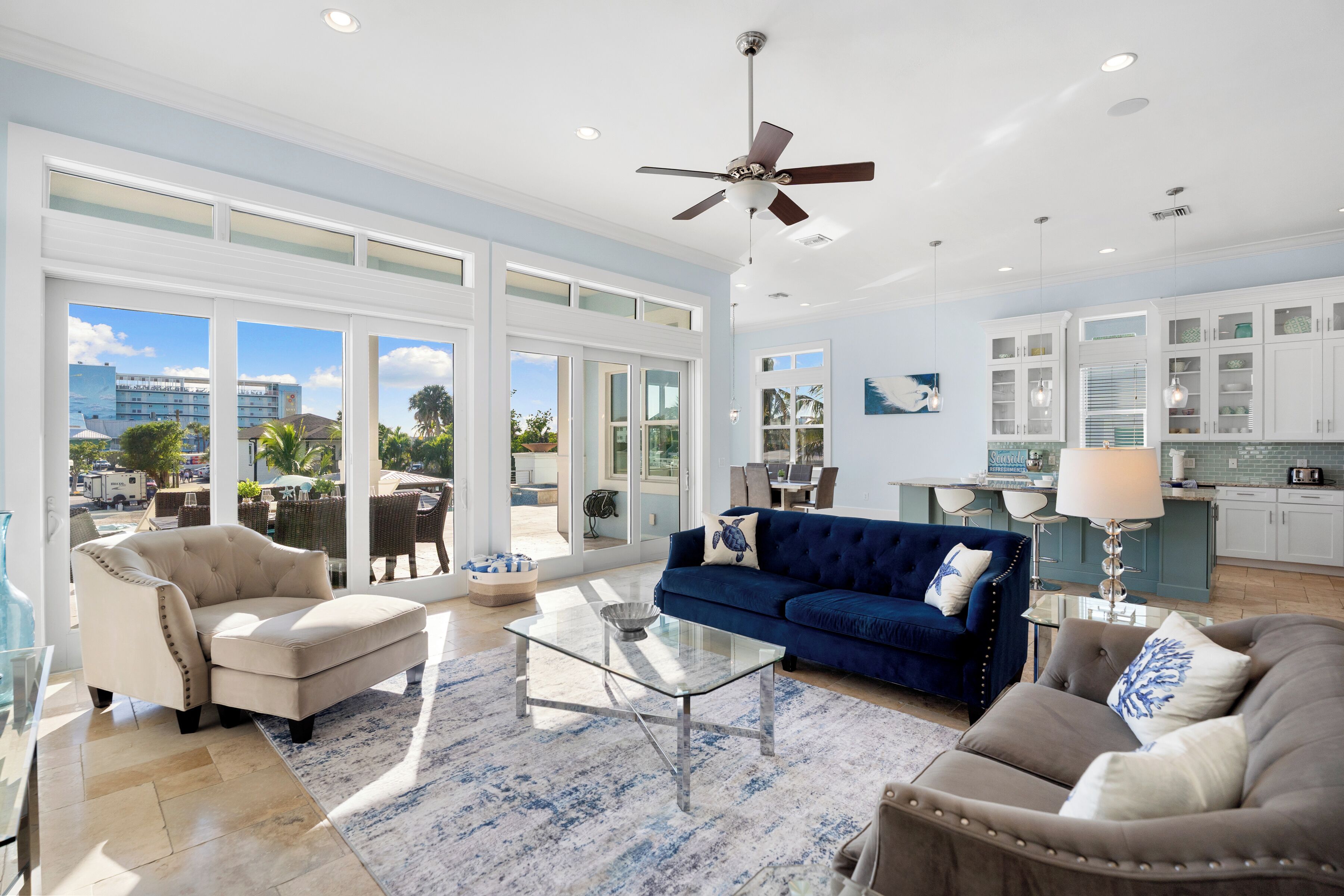 A spacious living room with a large glass coffee table, a blue sofa, and a beige armchair. There is a ceiling fan, a kitchen area with white cabinets, and large windows offering a view of the outdoors.