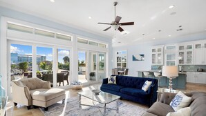 A spacious living room with a large glass coffee table, a blue sofa, and a beige armchair. There is a ceiling fan, a kitchen area with white cabinets, and large windows offering a view of the outdoors. - Seaside Villa Aqua luxury 4-bedroom villa in Fort Myers Beach incredible view (Fort Myers Beach)
