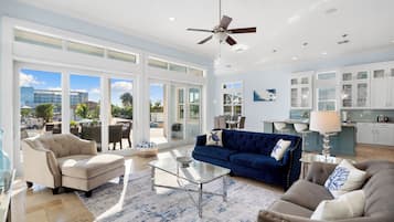 A spacious living room with a large glass coffee table, a blue sofa, and a beige armchair. There is a ceiling fan, a kitchen area with white cabinets, and large windows offering a view of the outdoors.