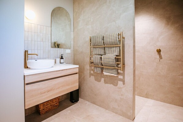 A modern bathroom with a white sink, gold faucet, and a mirror above it. There is a towel rack with checkered towels and a wooden basket under the sink.