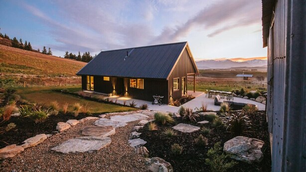 Una casa moderna con techo oscuro, un camino de piedra que lleva a la entrada y una vista de montañas al fondo.