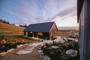 A modern house with a dark roof, a stone pathway leading to the entrance, and a view of mountains in the background. - Ranger`s Retreat | Mountain Views | Secluded (Lake Hāwea)