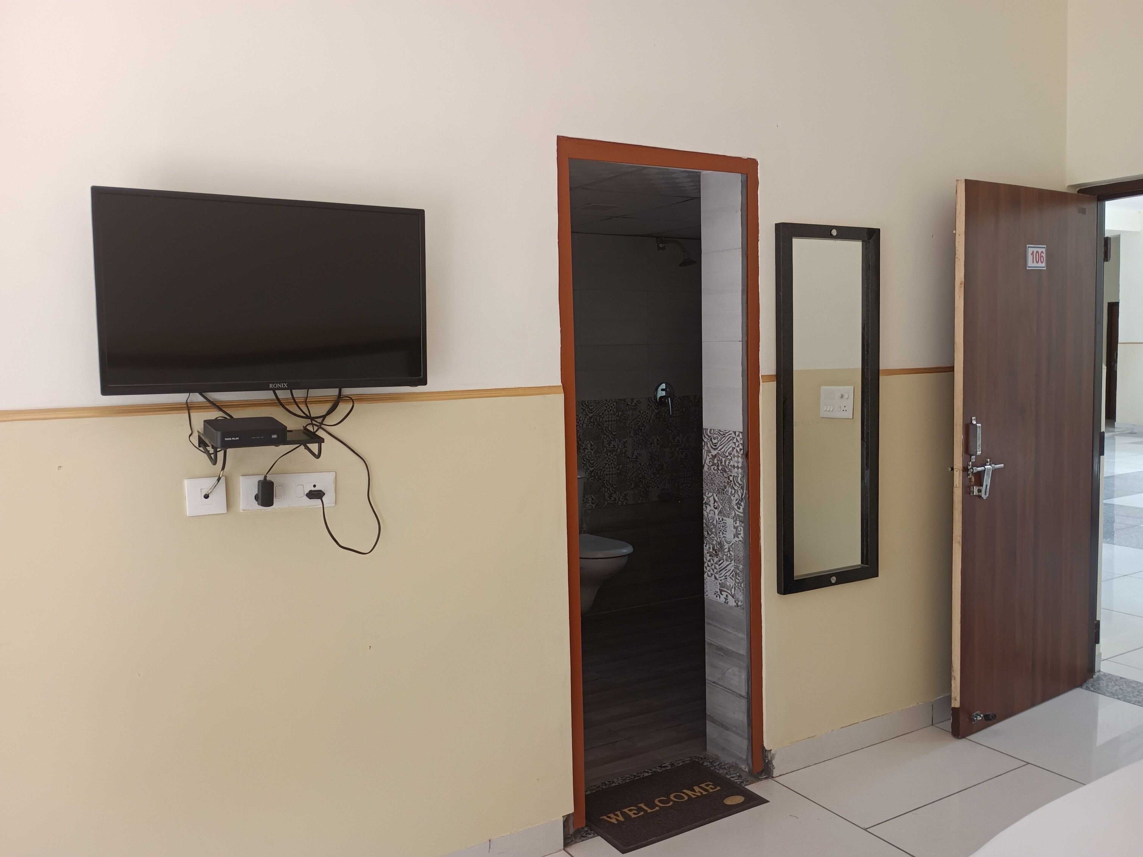 A room with a flat-screen TV mounted on the wall, a door with a 'WELCOME' mat, and a tiled floor.