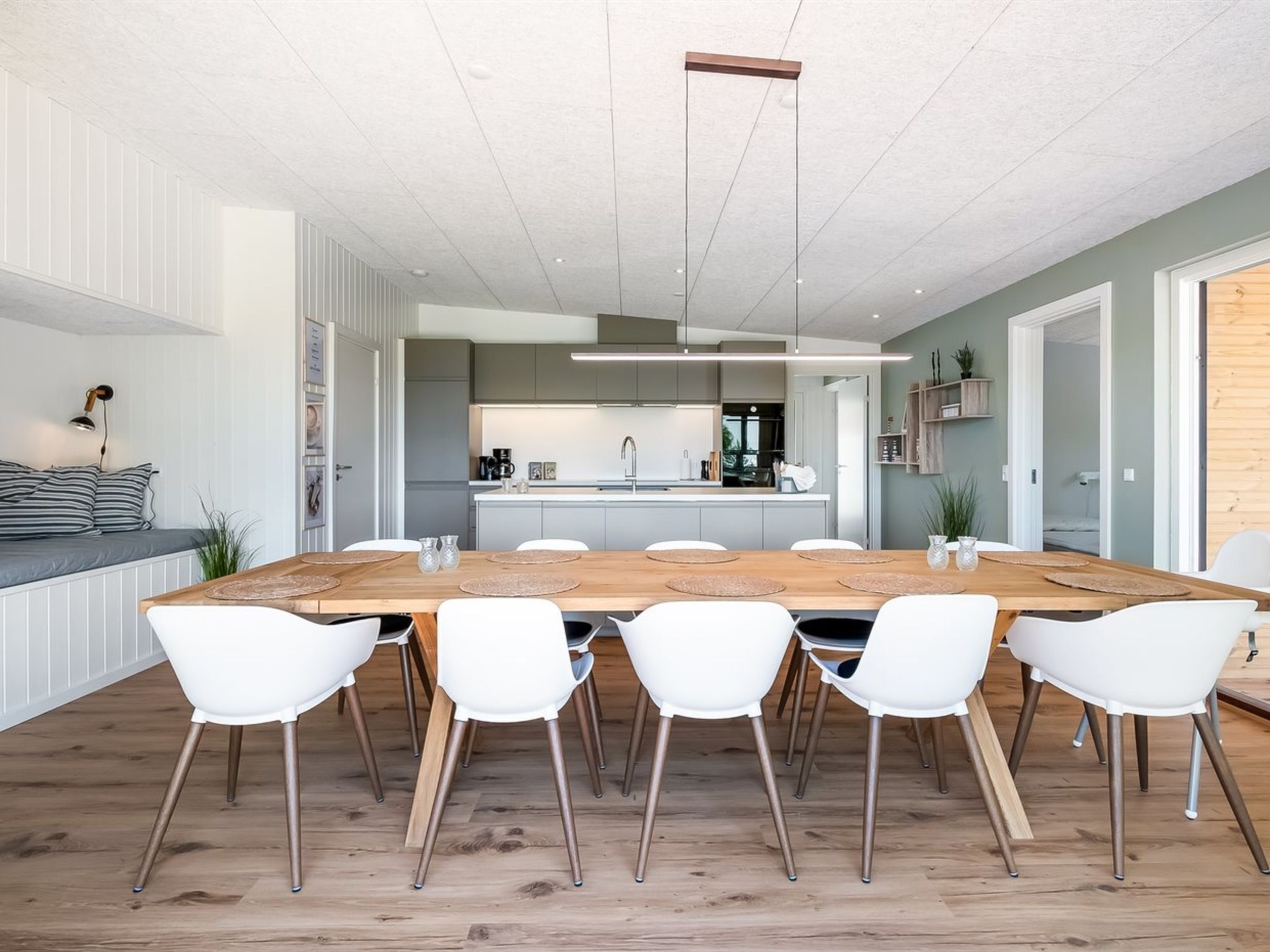 Un espace repas moderne avec une table en bois et des chaises blanches, une cuisine en arrière-plan et une vue sur l’extérieur.