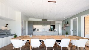 A modern dining area with a wooden table and white chairs, a kitchen in the background, and a view of the outdoors.