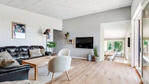 A modern living room with a black leather sofa, a white armchair, and a wooden coffee table. A flat-screen TV is mounted on the wall, and there is a dining area visible through an open door.