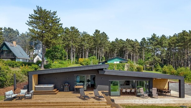A modern house with a large deck, outdoor seating, and a view of the surrounding forest.