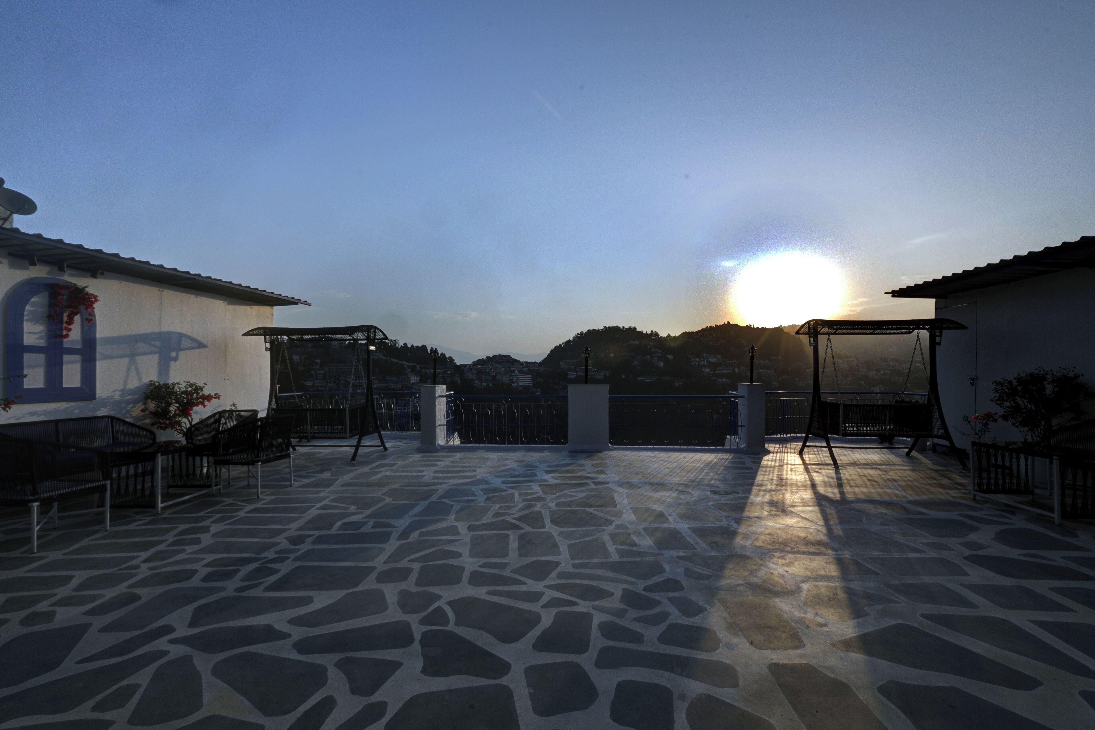 A courtyard with stone flooring, a pergola, and a view of the sun setting behind hills.