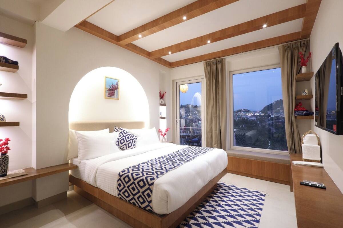 Une chambre d’hôtel moderne dotée d’un grand lit, d’une vue sur la ville, de poutres apparentes en bois au plafond et d’un téléviseur à écran plat.