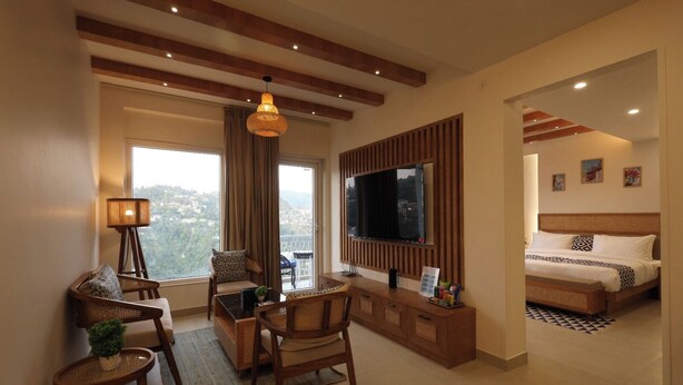 A modern living room with a large TV, comfortable seating, a balcony with a view, and a separate bedroom with a bed and artwork.