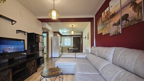 A living room with a large flat-screen TV, a beige sofa, and a dining area in the background.