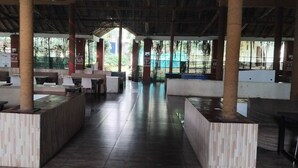 An indoor dining area with wooden tables and chairs, a central walkway, and large windows allowing natural light.