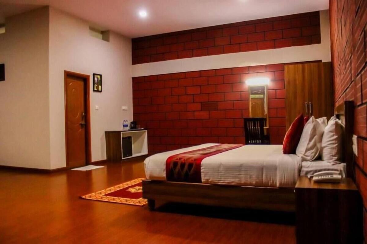 A hotel room with a brick accent wall, a bed with white bedding and a red throw, a wooden bench, and a small desk with a chair.