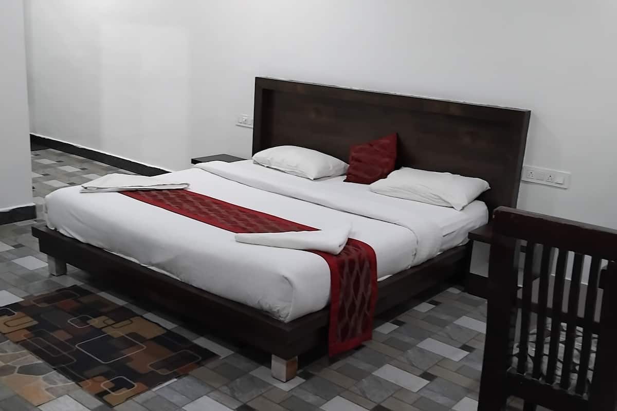 A neatly made bed with white bedding and a red throw, a wooden nightstand with a lamp, and a patterned rug on a tiled floor.