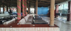 A spacious indoor area with long tables and benches, large pillars, and a stage with a blue backdrop. - Pavanputra Resort Hassan (Hassan)
