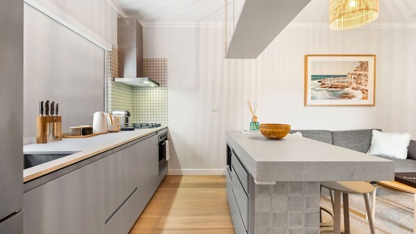 A modern kitchen with a central island, a dining area with a sofa, and a framed coastal landscape picture on the wall.