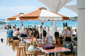 Un espace extérieur aménagé avec des tables et des chaises, une partie couverte avec des parasols, et une vue sur la plage et l’océan.