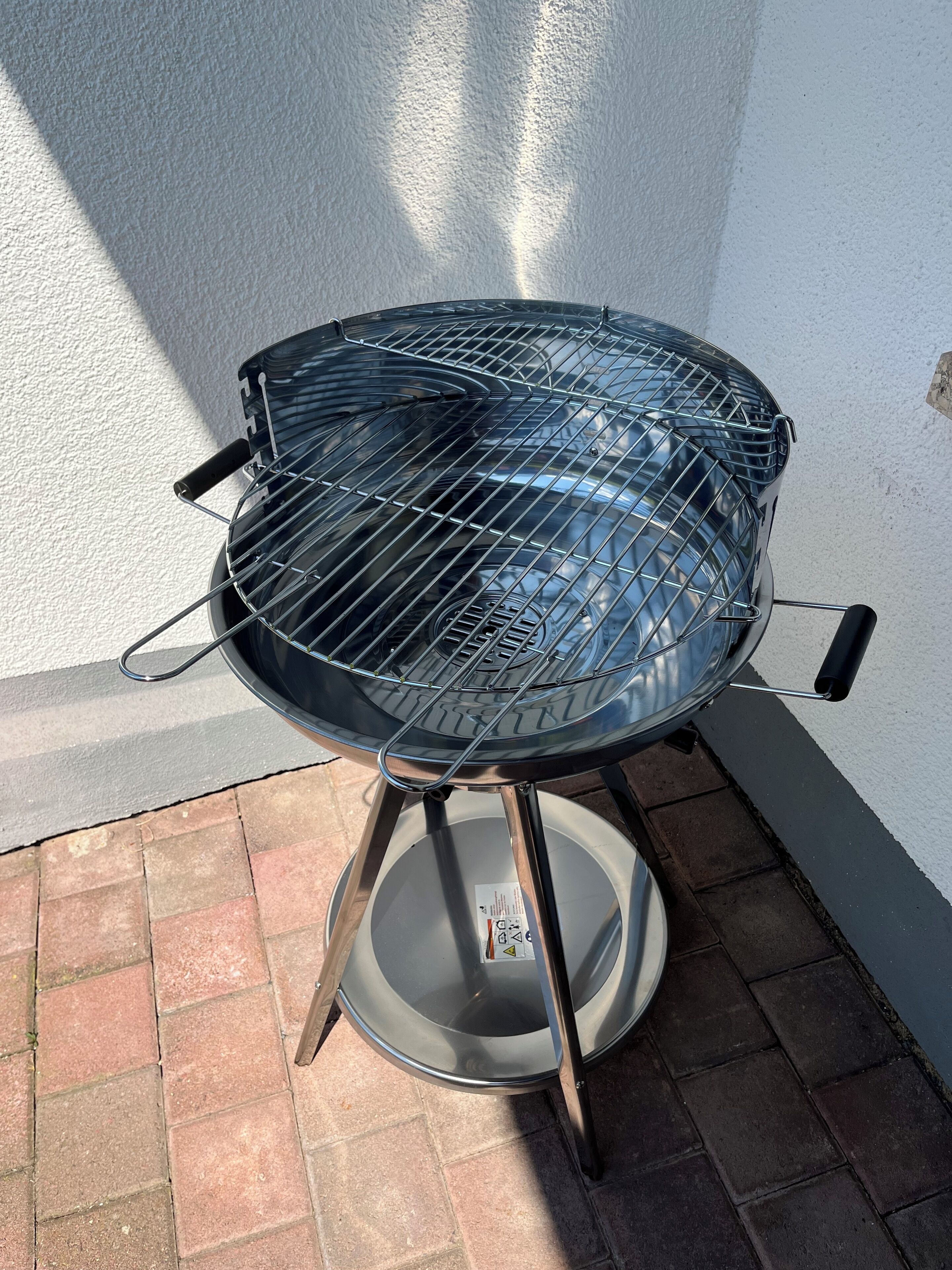 A round, metal barbecue grill with a wire grate, mounted on a metal stand with a circular base, positioned on a brick patio against a white wall.