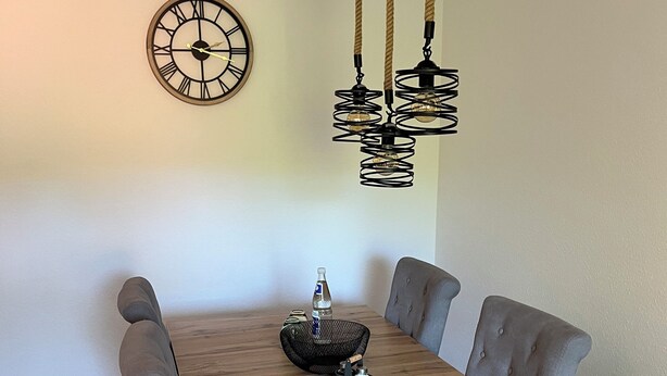 Comedor con mesa de madera, sillas tapizadas en gris, un reloj de pared y una lámpara de techo original con luces de cuerda.