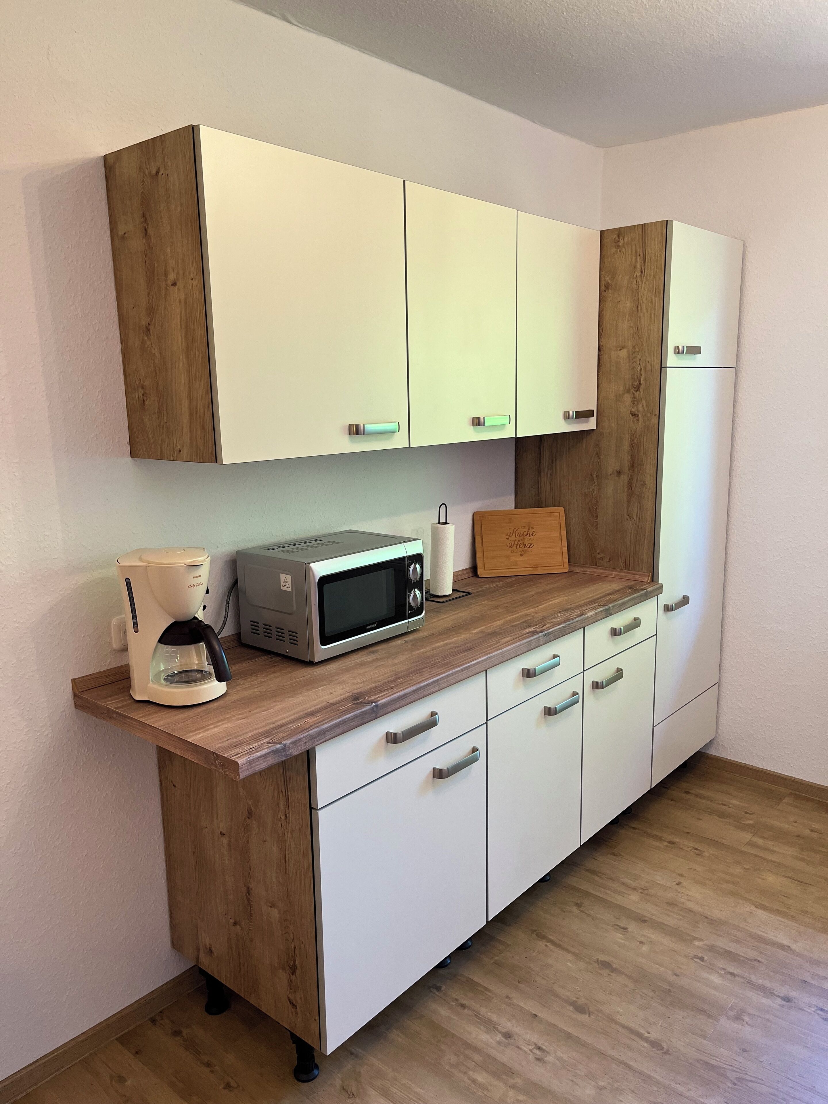 A compact kitchenette with white cabinets, a wooden countertop, a coffee maker, a microwave, and a cutting board.