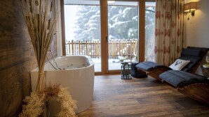 Ein Wellnessraum mit einer großen Badewanne, einer Couch und Blick auf eine verschneite Terrasse.