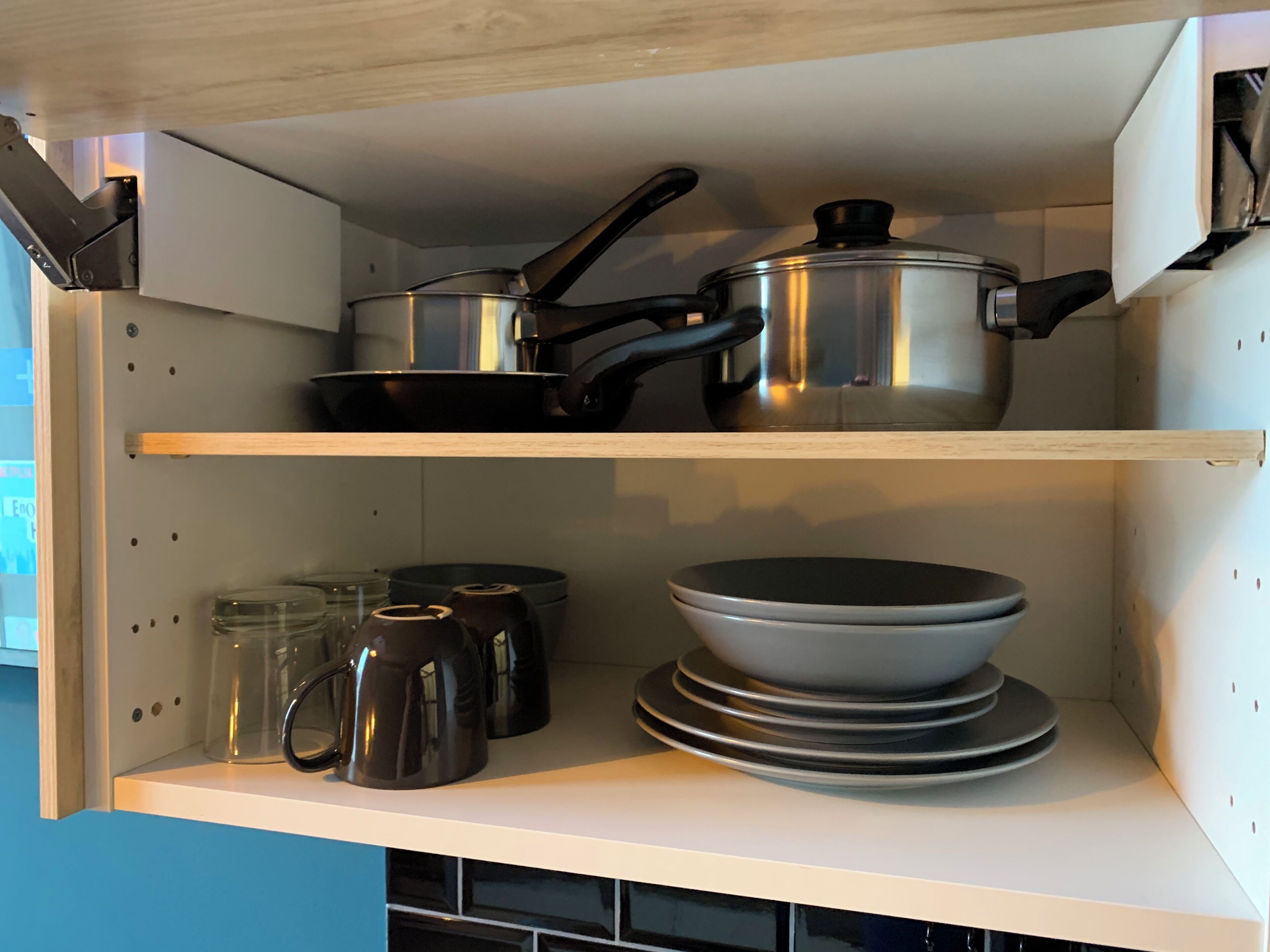 Ett kök med två hyllor: på den övre hyllan står en kastrull och en stekpanna i rostfritt stål, och på den nedre hyllan finns en stapel tallrikar och en kaffebryggare.