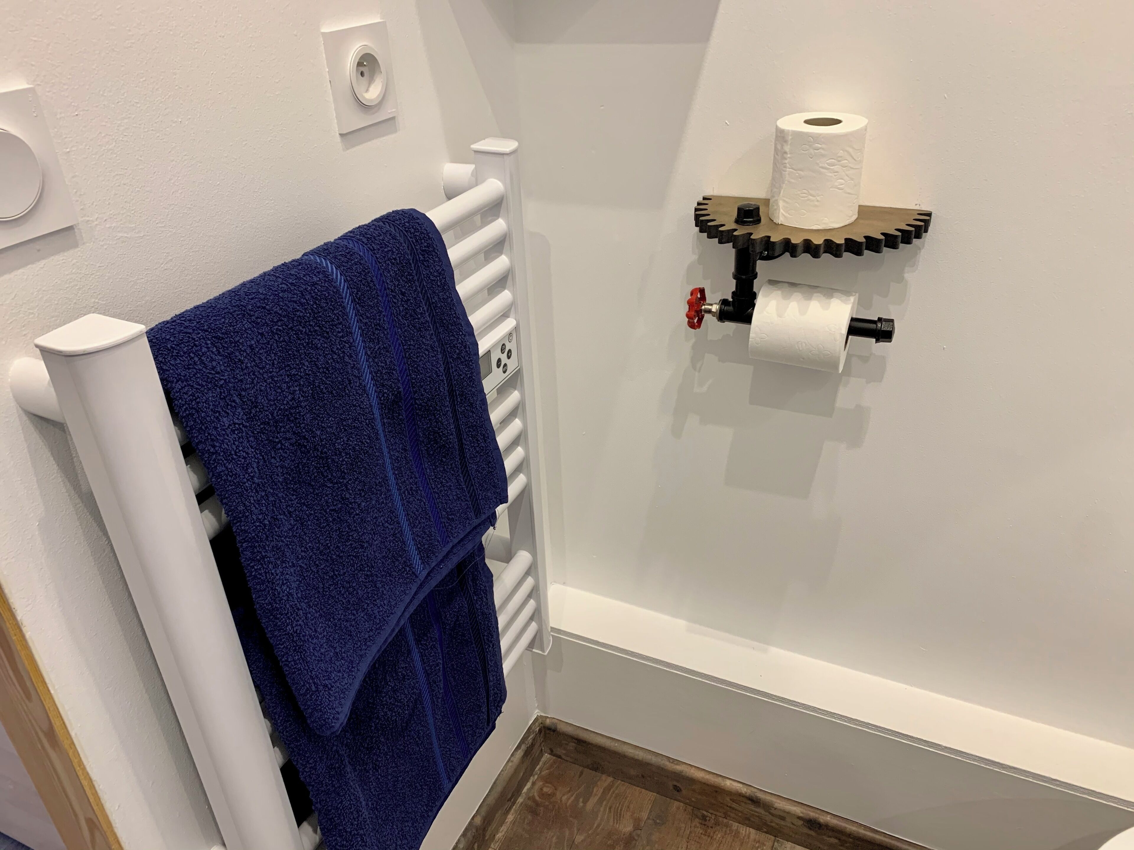 Une salle de bain avec un radiateur blanc, une serviette bleue suspendue à un porte-serviettes blanc, et un porte-rouleau de papier toilette avec un rouleau posé sur une étagère.
