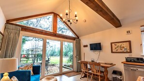 A room with a large sliding glass door, a dining table with chairs, a sofa, a TV mounted on the wall, and a kitchen area with a dishwasher. - Lodge in Soberton Near South Downs Riding (Southampton)