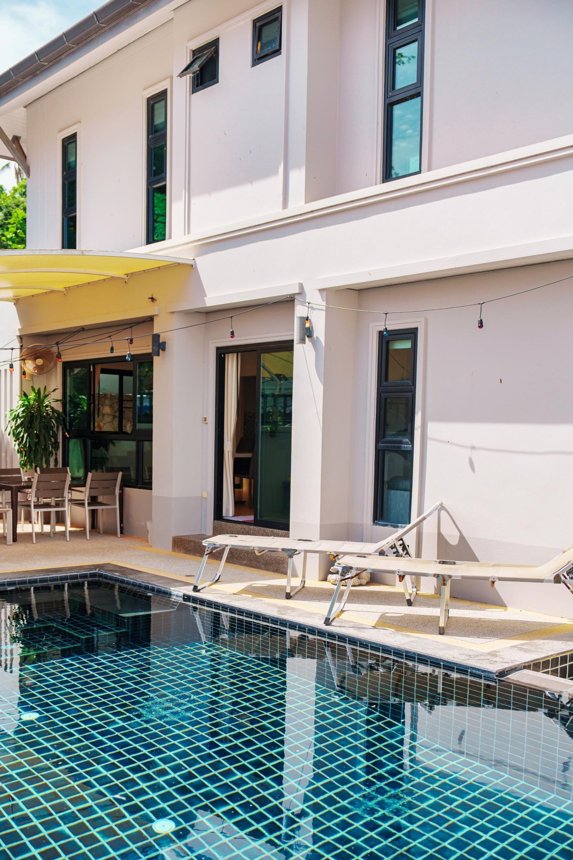 Una piscina con borde de azulejos, un porche cubierto con sillas y un edificio con amplias ventanas y una puerta corrediza.