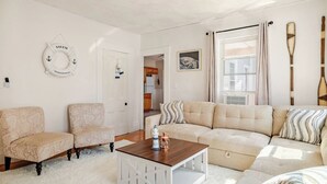 A living room with a beige sofa, two armchairs, a wooden coffee table, and a white door with a 'SALEM MASSACHUSETTS' sign. - Witch City Retreat w/ Tiki Bar (Salem)