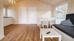 Mökki, jonka sisäseinät ovat puuta, ruokailutila valkoisilla huonekaluilla, kuviollinen koristetyyny sohvan päällä ja kaukosäädin pöydällä.