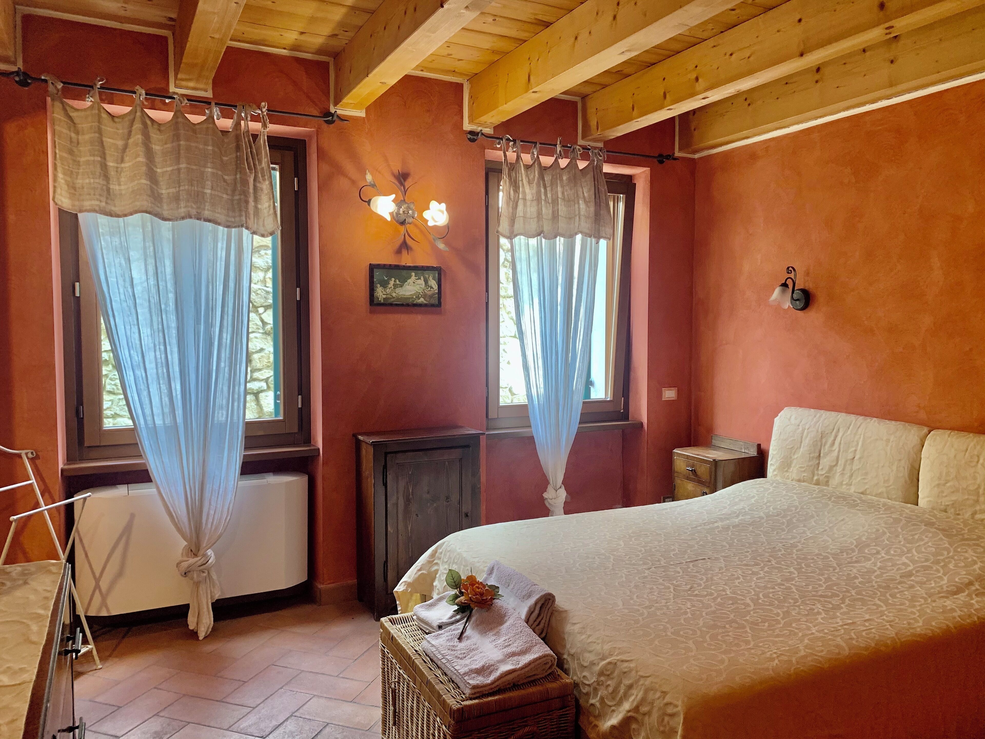 Un dormitorio con una cama, dos ventanas con cortinas translúcidas, una lámpara de pared, un radiador y una mesita de noche pequeña.